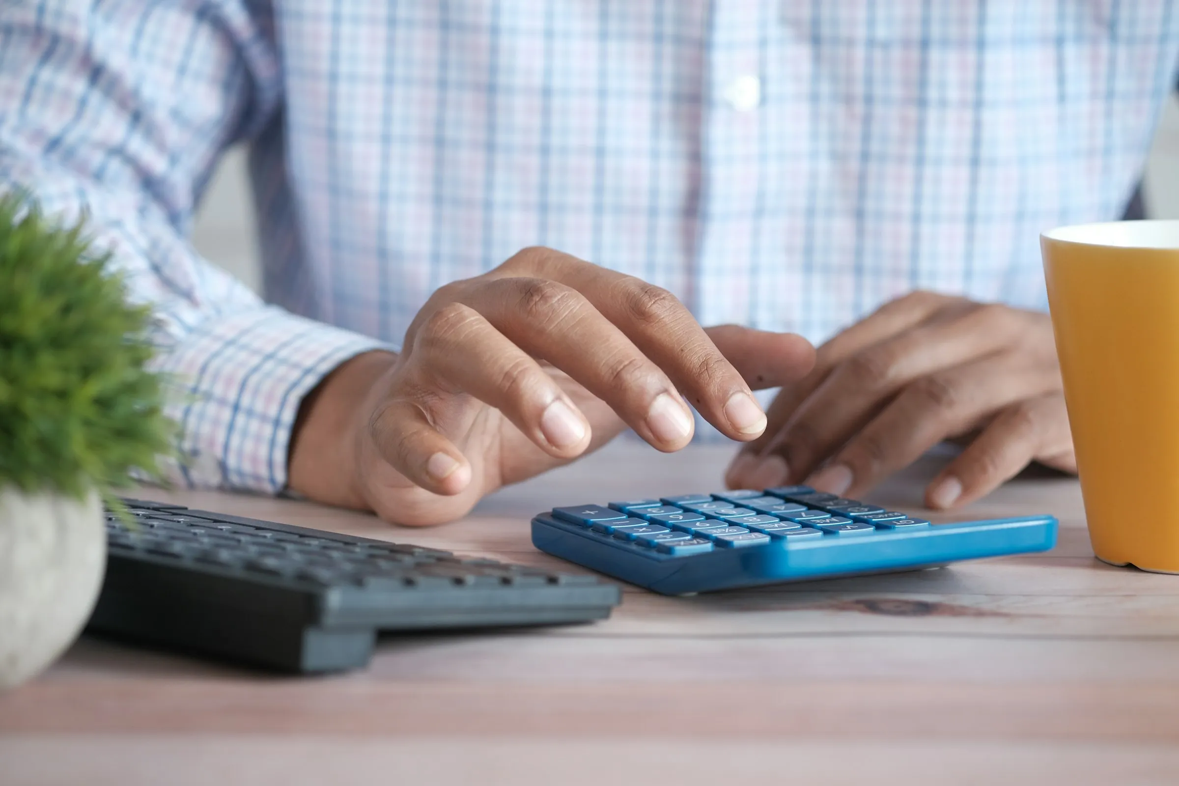 Hands working a calculator at a desk beside a keyboard and a mug